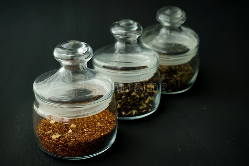 Glass clear jars round bellied low for tea with lid on black textured background with custard black and green tea inside stock image