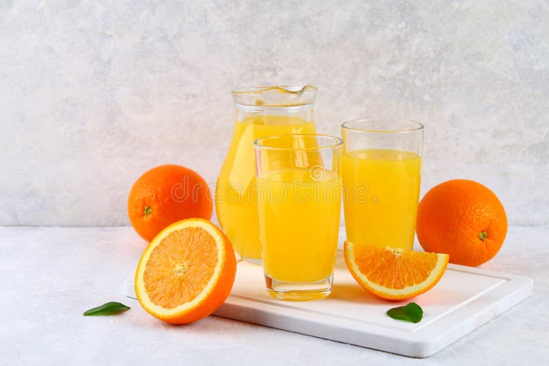 Glass cups and a pitcher of fresh orange juice with slices of orange and yellow tubes on a light gray table.