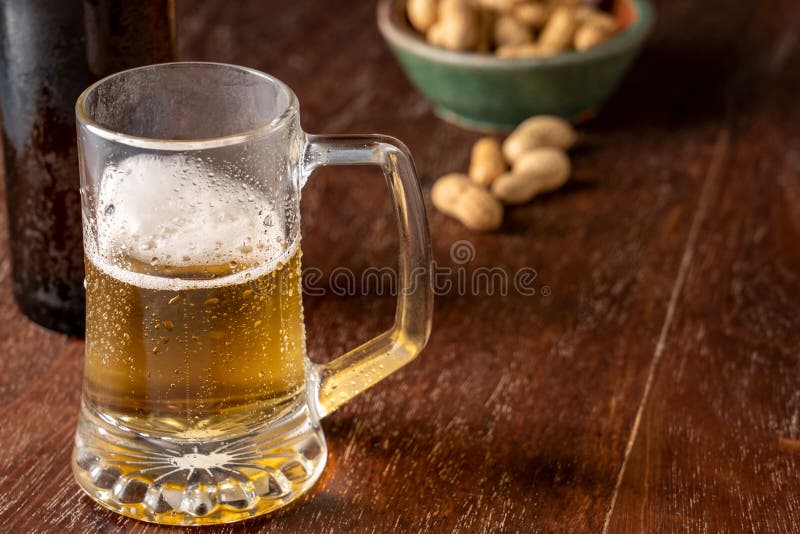 A glass of freshly poured beer on a wooden table next to some nuts as a snack