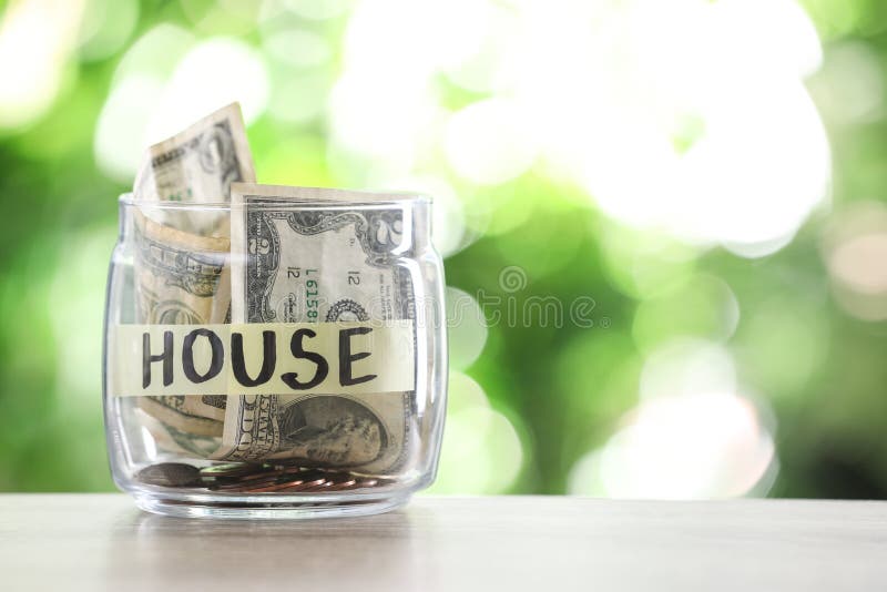 Glass jar with money and label HOUSE on table against blurred background