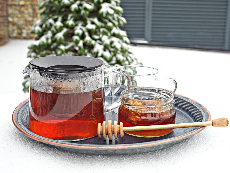 Glass kettle of hot tea and jar of honey on table covered by snow