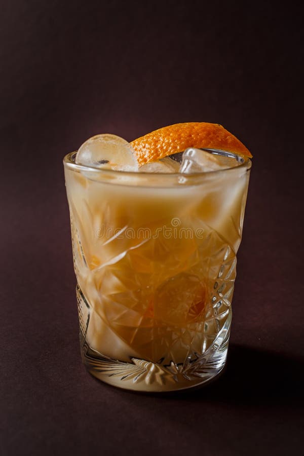 Glass of orange alcohol cocktail with ice and slice of orange on elegant dark brown background.