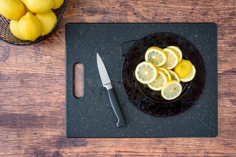 Glass plate with fresh lemon slices, and paring knife, on a black cutting board, wood table, basket of lemons