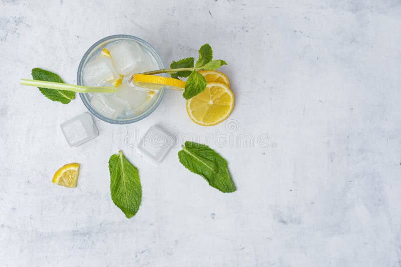 A glass of refreshing lemon water with ice cubes, mint leaves and a straw stands on a light marble background. A citrus drink for stock photo