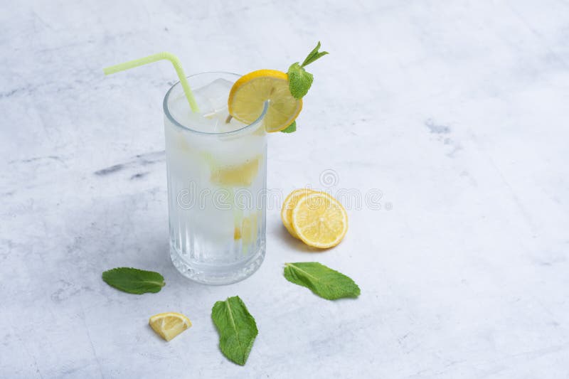 A glass of refreshing lemon water with ice cubes, mint leaves and a straw stands on a light marble background. A citrus drink for stock image
