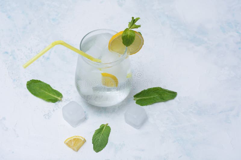 A glass of refreshing lemon water with ice cubes, mint leaves and a straw stands on a light marble background. A citrus drink for stock photos