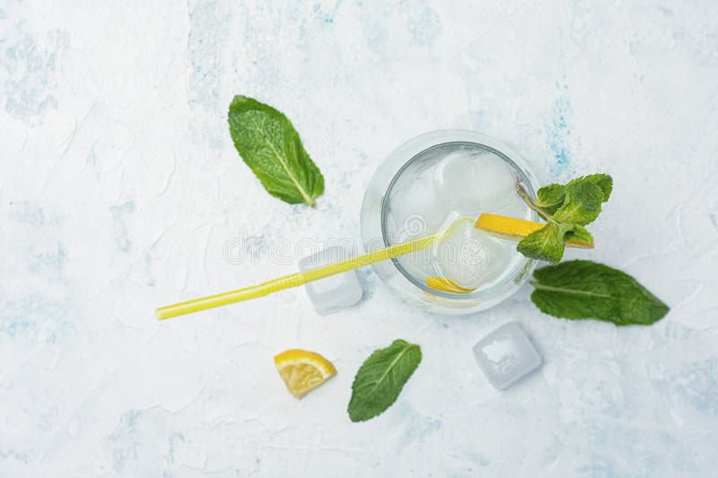 A glass of refreshing lemon water with ice cubes, mint leaves and a straw stands on a light marble background. A citrus drink for stock photo