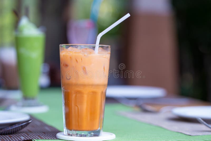 A glass of Thai iced tea with white straw put on dining table with green tea in a restaurant background.