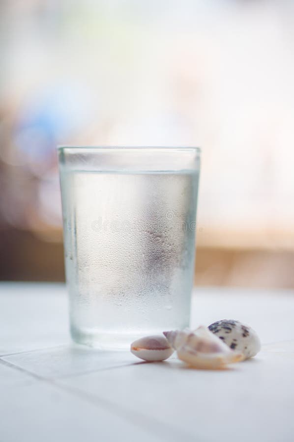 Glass of water with drops and small shells on pool side in tropical beach resort