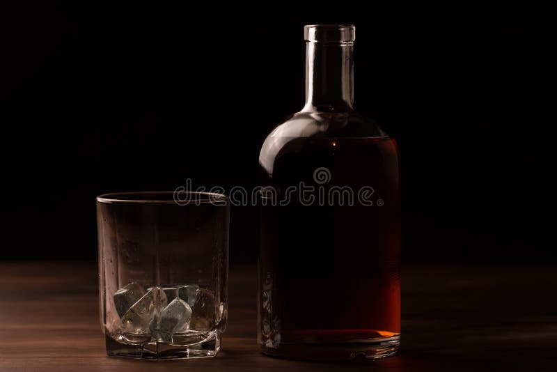 Glass of whiskey with ice and a bottle on a wooden table.