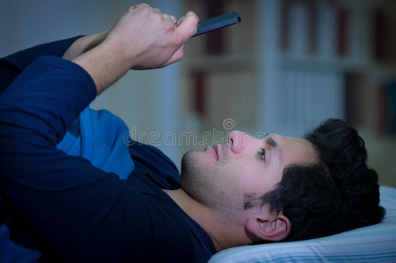 Gli uomini sociali della persona dedita di media sul letto non dormono perché Smart Phone del gioco, fondo della stanza immagini stock libere da diritti Gli uomini sociali della persona dedita di media sul letto non dormono perché Smart Phone del gioco, fondo della stanza immagini stock libere da diritti