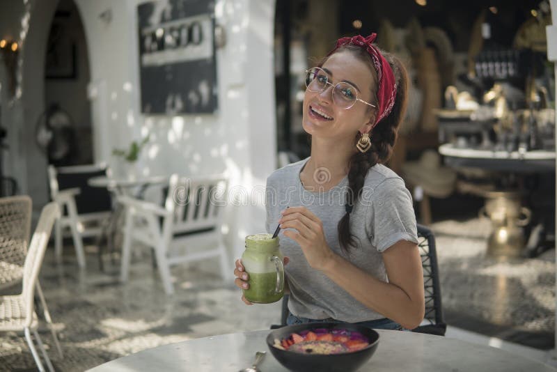 Glimlachend donkerbruin meisje in een rode bandana en met vlecht die een tropische lunch in open koffie, groene cocktail en kom h royalty-vrije stock afbeelding Glimlachend donkerbruin meisje in een rode bandana en met vlecht die een tropische lunch in open koffie, groene cocktail en kom h royalty-vrije stock afbeelding