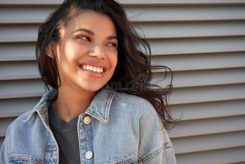 Smiling young African American teen girl looking away and laughing standing outdoor, headshot. Happy stylish millennial mixed race woman wearing denim jacket with dental smile closeup face portrait. Smiling young African American teen girl looking away and laughing standing outdoor, headshot. Happy stylish millennial mixed race woman wearing denim jacket with dental smile closeup face portrait.