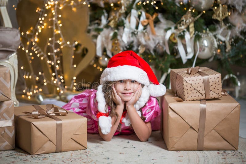 Glimlachend leuk meisje dichtbij met santahoed dichtbij giften en Kerstboom Nieuwe jaar of Kerstmisviering thuis royalty-vrije stock foto Glimlachend leuk meisje dichtbij met santahoed dichtbij giften en Kerstboom Nieuwe jaar of Kerstmisviering thuis royalty-vrije stock foto