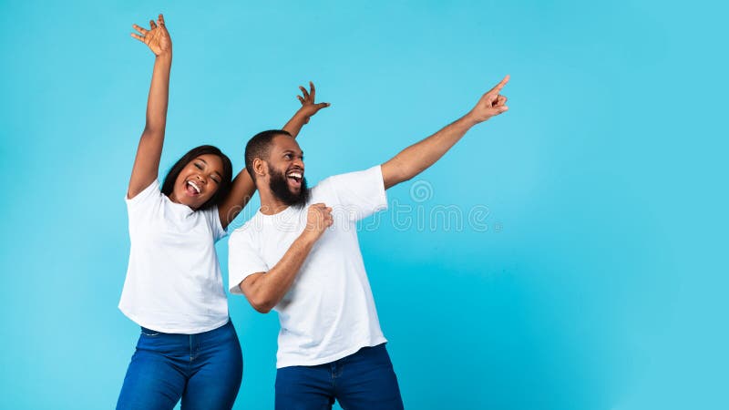 Cool Offer Concept. Portrait Of Excited Black Young Couple Having Fun, Celebrating Victory, Happy Woman Raising Hands Up, Man Pointing Finger Aside At Copy Space On Blue Studio Background. Cool Offer Concept. Portrait Of Excited Black Young Couple Having Fun, Celebrating Victory, Happy Woman Raising Hands Up, Man Pointing Finger Aside At Copy Space On Blue Studio Background