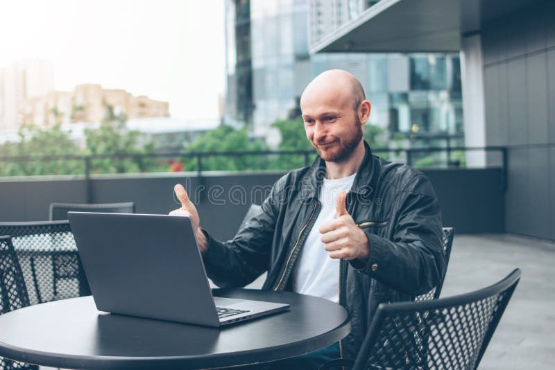 Glimlachende aantrekkelijke volwassen succesvolle kale gebaarde mens in zwart jasje met laptop in straatkoffie bij stad stock fotografie Glimlachende aantrekkelijke volwassen succesvolle kale gebaarde mens in zwart jasje met laptop in straatkoffie bij stad stock fotografie