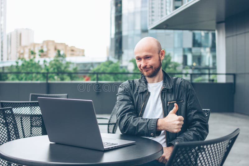 Glimlachende aantrekkelijke volwassen succesvolle kale gebaarde mens in zwart jasje met laptop in straatkoffie bij stad royalty-vrije stock afbeeldingen Glimlachende aantrekkelijke volwassen succesvolle kale gebaarde mens in zwart jasje met laptop in straatkoffie bij stad royalty-vrije stock afbeeldingen