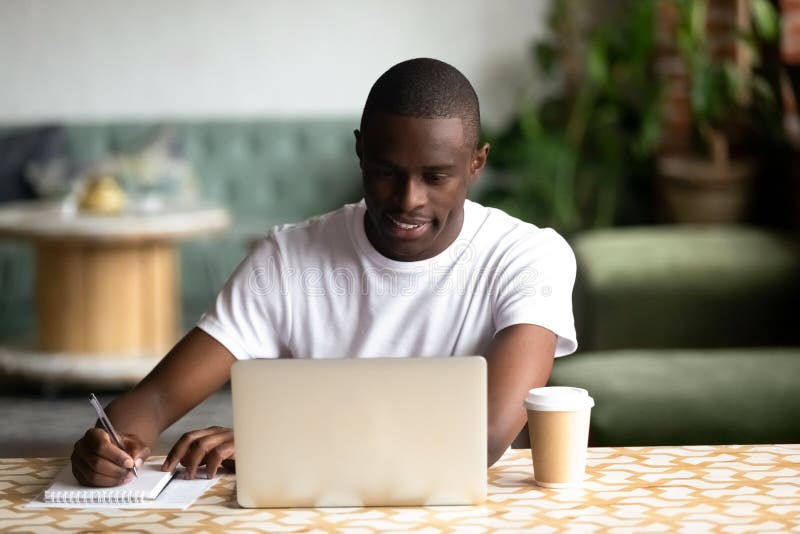 Smiling focused African American man using laptop, studying or working in coffee house, looking at screen, making notes, student preparing for test or exam in cafe, writing information in notebook. Smiling focused African American man using laptop, studying or working in coffee house, looking at screen, making notes, student preparing for test or exam in cafe, writing information in notebook