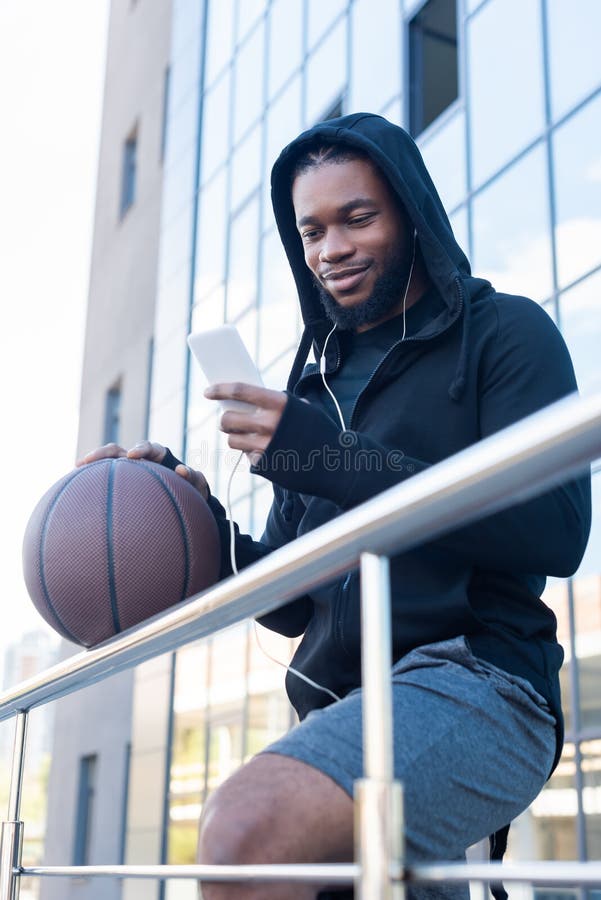 glimlachende Afrikaanse Amerikaanse mens in oortelefoons die smartphone gebruiken terwijl het houden van basketbalbal stock afbeelding glimlachende Afrikaanse Amerikaanse mens in oortelefoons die smartphone gebruiken terwijl het houden van basketbalbal stock afbeelding