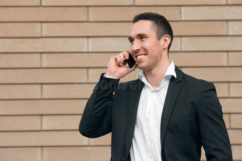 Glimlachende gelukkige mens die op mobiele telefoon in een zwart kostuum spreken, gelukkige moderne mens outdoors stock afbeeldingen Glimlachende gelukkige mens die op mobiele telefoon in een zwart kostuum spreken, gelukkige moderne mens outdoors stock afbeeldingen