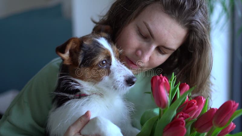 Glimlachende kaukasische vrouw met een bouquet tulpen die haar hond kussen die thuis op de grond zit. lifestyle