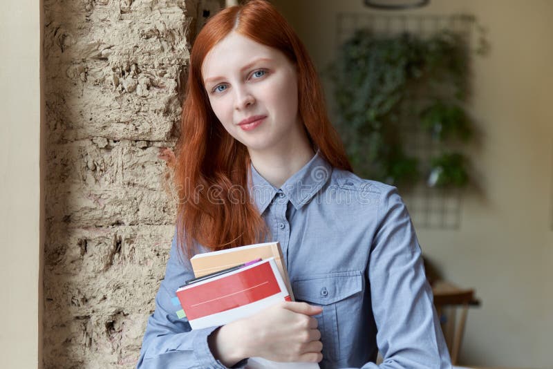 Glimlachende leuke schuwe jonge vrouw met lang rood haar die en boeken bevinden zich houden royalty-vrije stock foto Glimlachende leuke schuwe jonge vrouw met lang rood haar die en boeken bevinden zich houden royalty-vrije stock foto