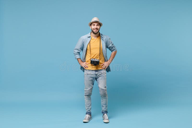 Smiling traveler tourist man in yellow summer clothes with photo camera isolated on blue background. Male passenger traveling abroad on weekends. Air flight journey. Stand with arms akimbo on waist. Smiling traveler tourist man in yellow summer clothes with photo camera isolated on blue background. Male passenger traveling abroad on weekends. Air flight journey. Stand with arms akimbo on waist