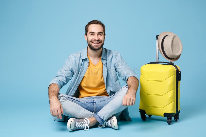 Smiling traveler tourist man in summer yellow casual clothes isolated on blue wall background. Male passenger traveling abroad to travel on weekend. Air flight journey concept. Sitting near suitcase. Smiling traveler tourist man in summer yellow casual clothes isolated on blue wall background. Male passenger traveling abroad to travel on weekend. Air flight journey concept. Sitting near suitcase