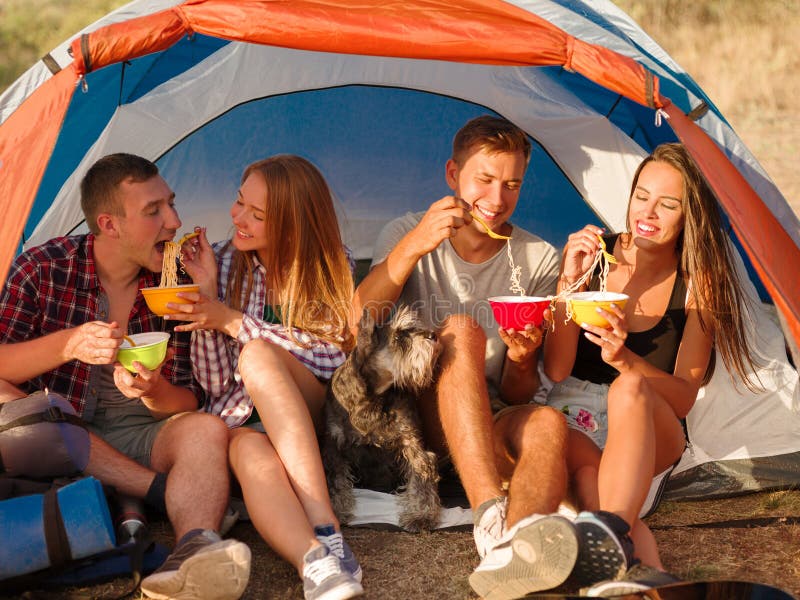 Glimlachende vrienden die snel voedselnoedels op een het kamperen reis eten Wandelaars die op een tentachtergrond eten Actief vak stock foto's Glimlachende vrienden die snel voedselnoedels op een het kamperen reis eten Wandelaars die op een tentachtergrond eten Actief vak stock foto's