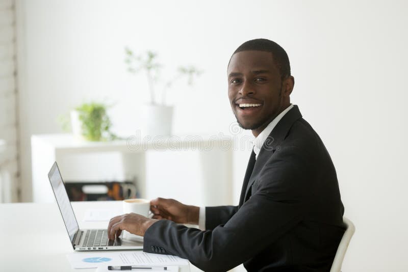 Smiling positive African American looking at camera working at laptop and drinking coffee. Black company financial manager preparing report using computer. Friendly employee posing in small office. Smiling positive African American looking at camera working at laptop and drinking coffee. Black company financial manager preparing report using computer. Friendly employee posing in small office
