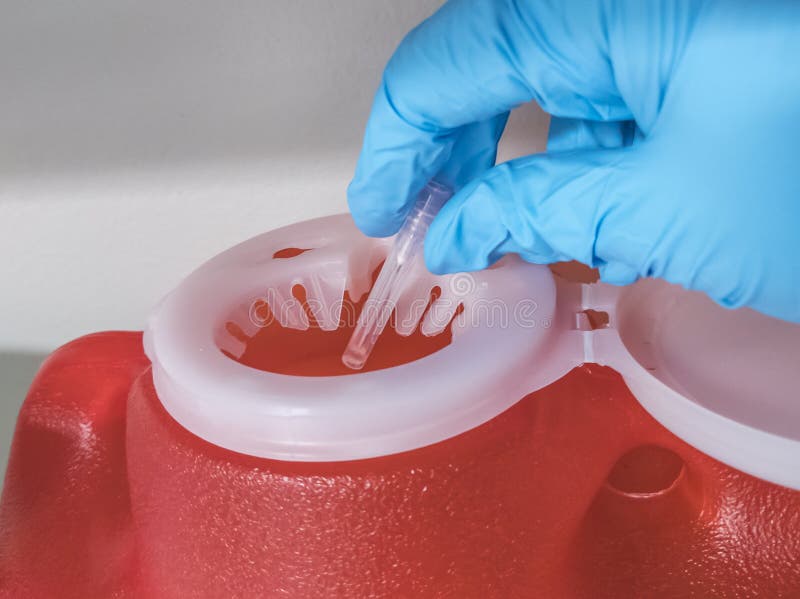 Gloved hand dropping a used needle into a bright red sharps container