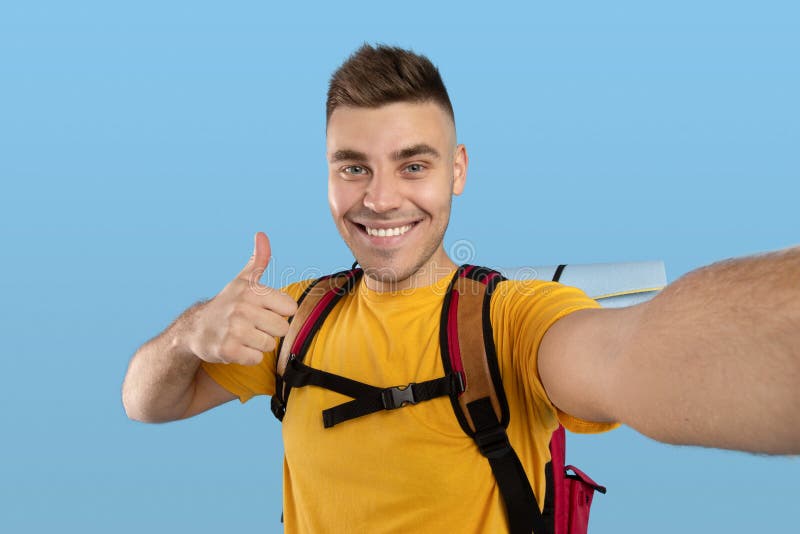 Glücklicher männlicher Reisender mit Rucksack mit Daumen-up Geste, während er sich über blauem Studio-Hintergrund selfie stockfotografie