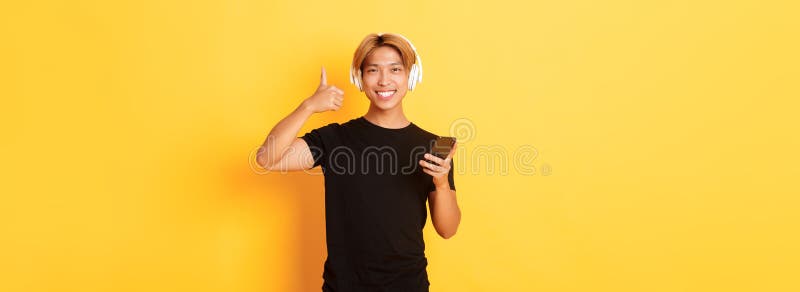 Glücklich zufriedener asiatischer Typ mag Musik oder Podcast zeigen Daumen in der Genehmigung halten Handy gelb stehen lizenzfreies stockfoto