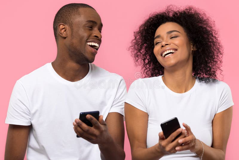 Glückliche afrikanische Paare, die unter Verwendung der Smartphones lokalisiert auf rosa Hintergrund lachen stockfotos