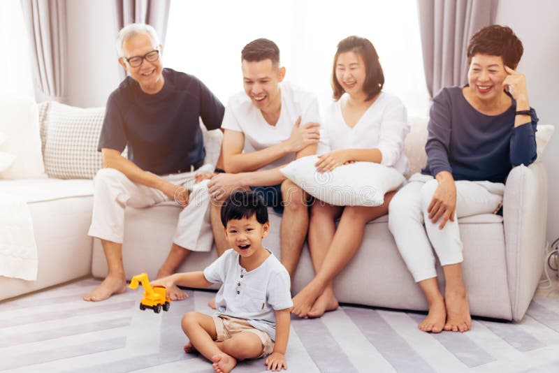 Glückliche asiatische Großfamilie, die zusammen auf Sofa und dem aufpassenden kleinen Kind spielt Spielzeug auf dem Boden mit Glü lizenzfreie stockbilder Glückliche asiatische Großfamilie, die zusammen auf Sofa und dem aufpassenden kleinen Kind spielt Spielzeug auf dem Boden mit Glü lizenzfreie stockbilder