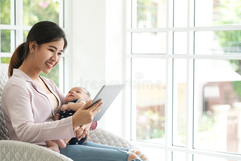 Glückliche attraktive asiatische Mutter mit dem Kind, das an der Couch im Wohnzimmer sucht Elternbildung im Sozialen Netz sitzt lizenzfreie stockfotos Glückliche attraktive asiatische Mutter mit dem Kind, das an der Couch im Wohnzimmer sucht Elternbildung im Sozialen Netz sitzt lizenzfreie stockfotos