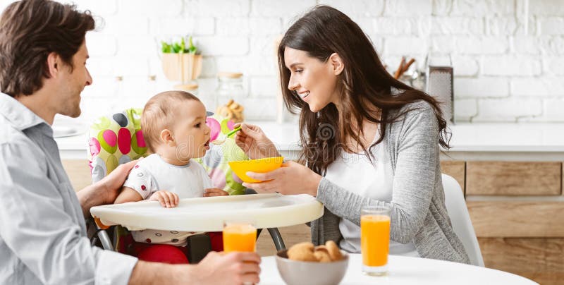 Glückliche Familie genießt gemeinsam Mahlzeit in Küche. Eine Familie um einen Tisch in ihrer Küche herum versammelt, um gemeinsam zu essen. eine Mutter füttert ihr Baby aus einer Schüssel mit einem grünen Löffel, während der Vater darauf schaut zu lächeln