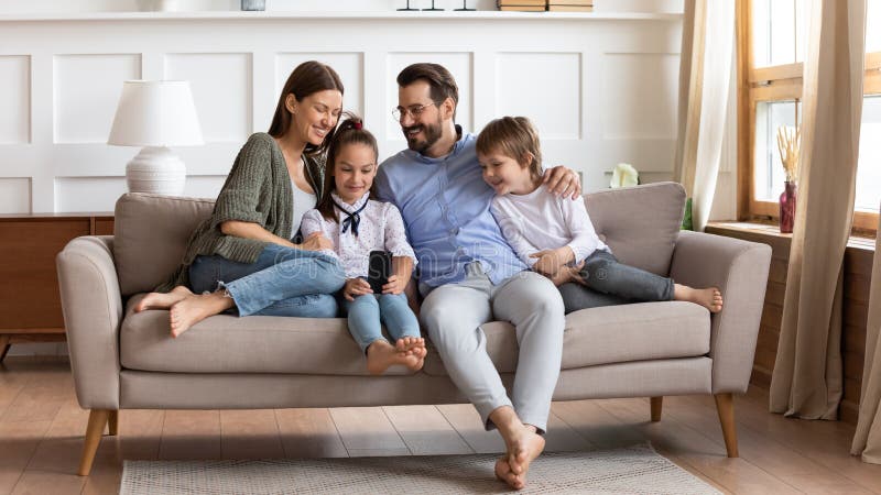 Happy young Caucasian family with little kids sit relax on comfortable sofa in living room using smartphone together, smiling parents rest with small children on couch at home browsing cellphone. Happy young Caucasian family with little kids sit relax on comfortable sofa in living room using smartphone together, smiling parents rest with small children on couch at home browsing cellphone