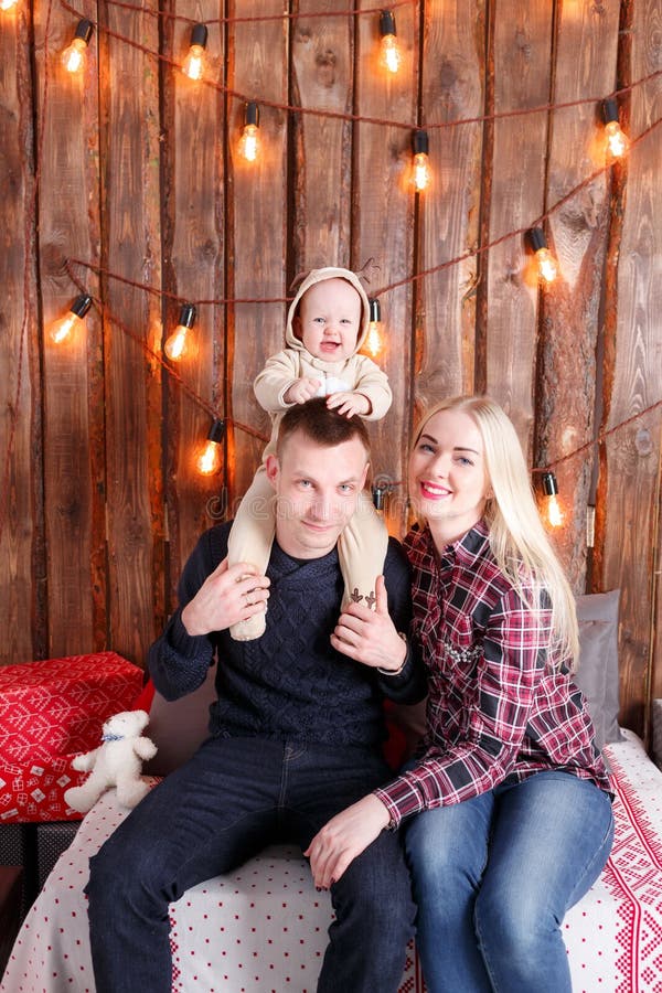 Glückliche Familie am Weihnachten Die Eltern und das Babysitzen Wand von hölzernen Planken und von Girlande lizenzfreie stockbilder Glückliche Familie am Weihnachten Die Eltern und das Babysitzen Wand von hölzernen Planken und von Girlande lizenzfreie stockbilder