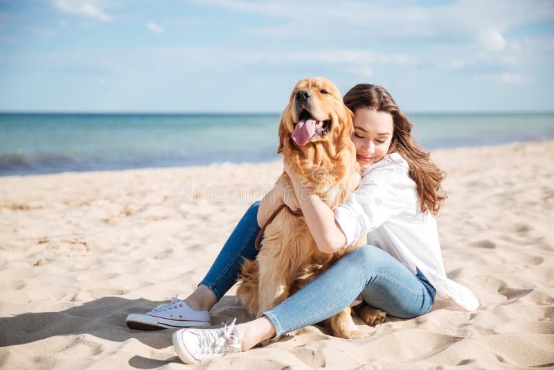 Glückliche Frau, die ihren Hund auf dem Strand sitzt und umarmt stockbild