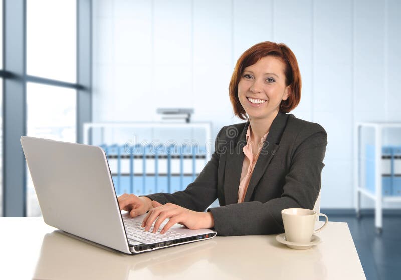 Glückliche Geschäftsfrau mit dem roten Haar lächelnd bei der Arbeit schreibend auf Computerlaptop am modernen Schreibtisch stockfotografie Glückliche Geschäftsfrau mit dem roten Haar lächelnd bei der Arbeit schreibend auf Computerlaptop am modernen Schreibtisch stockfotografie
