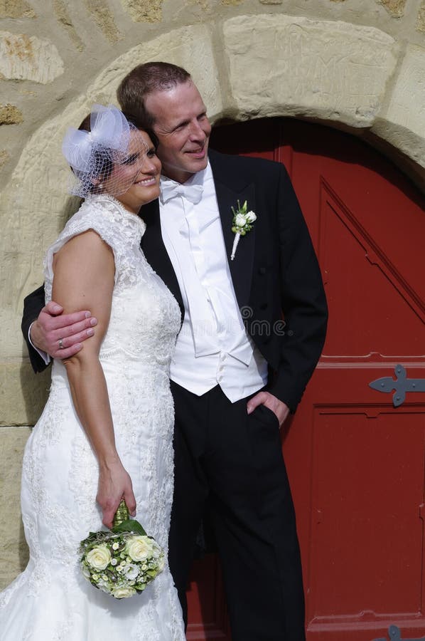 Caucasian wedding couple in vintage Dress in front of an old red door and natuarl stone wall of a historic building smiling, bride and groom in wihte wedding dress and black suit / smoking with bridal bouque looking to the side. Caucasian wedding couple in vintage Dress in front of an old red door and natuarl stone wall of a historic building smiling, bride and groom in wihte wedding dress and black suit / smoking with bridal bouque looking to the side