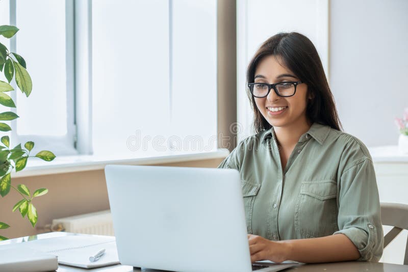 Glückliche indische junge Frau mithilfe des Büros der Laptop Arbeitsstudie zu Hause. Glückliche indische junge tragen Brille der erwachsenen Frau mithilfe der PC-Laptop-Computers, die das Amt zu Hause Prüfung auf den Tisch sitzt funktioniert. glücklicher weiblicher Berufsfreiberufler, der aufpassendes Webinar Online-Schulungen lernt