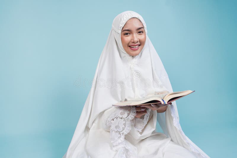 Glückliche junge asiatische Muslimfrau im Gebet, die sich hinsetzt und quran über isolierten blauen Hintergrund liest. Hadsch-Konz stockfotografie