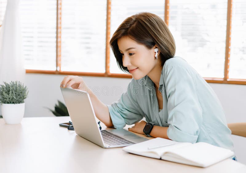 Glückliche junge schöne asiatische Frau mithilfe der Laptopfunktion entfernt und der drahtlosen Kopfhörer, die auf den Tisch in Ha stockfotografie