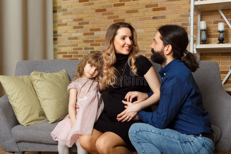 Glückliche junge Vatermutter und -tochter sitzen auf Weidensofa zu Hause. das Bild einer glücklichen Familie, die das zweite Kinderstudio erwartet