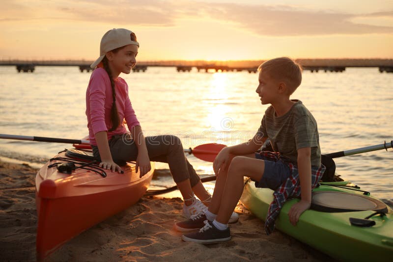 Glückliche Kinder, die auf Kajaks Nähe Fluss bei Sonnenuntergang sitzen. Sommercamp lizenzfreies stockfoto