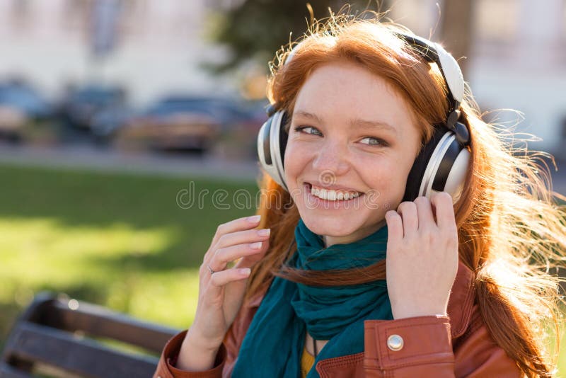 Portrait of happy smiling redhead young lady in bright scarf enjoying music on the bench in the park. Portrait of happy smiling redhead young lady in bright scarf enjoying music on the bench in the park