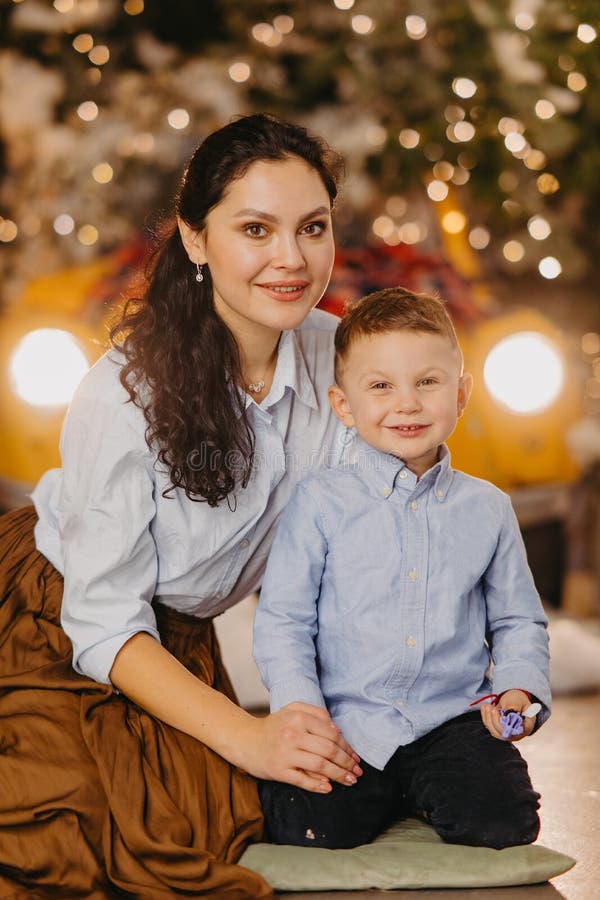 Glückliche Mutter mit Sohn sitzen auf dem Boden gegen Hintergrund der glühenden Scheinwerfern und Weihnachtsdekorationen stockfotografie