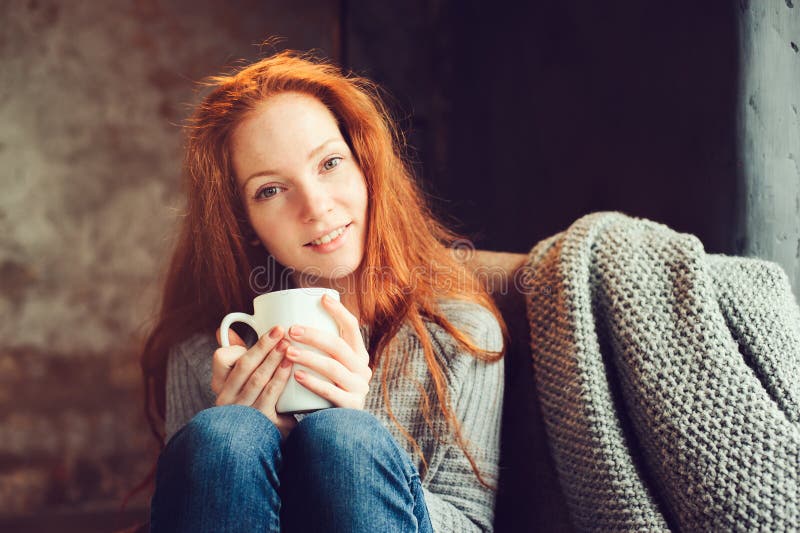 Glückliche Rothaarigefrau, die sich zu Hause im gemütlichen Winter- oder Herbstwochenende mit dem Buch und Schale heißem Tee, sit stockbild
